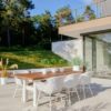 Silla de comedor Delphine Mocca ambiente, se ven seis sillas y una mesa de teca en una terraza muy bonita que da a una puerta acristalada por la derecha y a un bancal muy verde a la izquierda