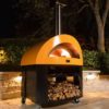 Base Horno 5 pizze Híbrido Amarillo con base. Colocado en una terraza de piedra con una jardinera detrás. Es un ambiente nocturno y la base del horno está llena de leña.