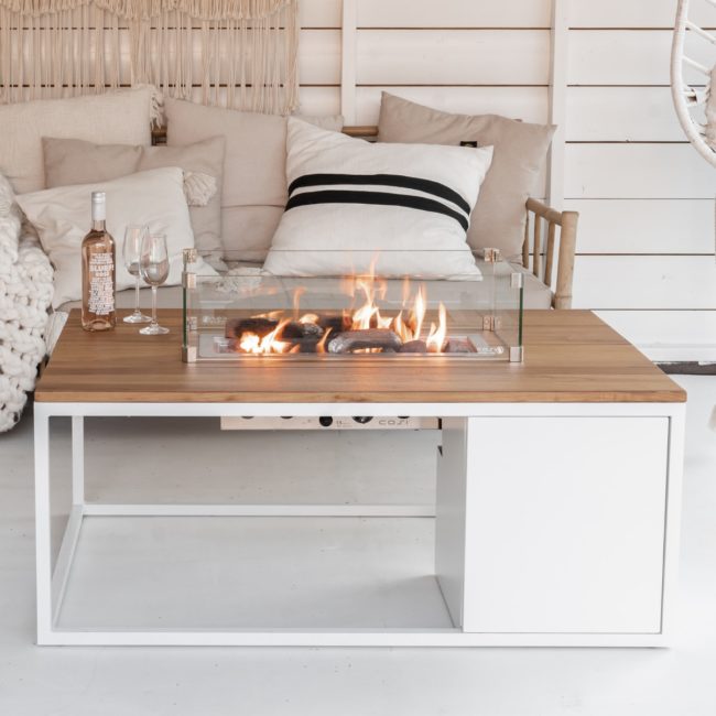 Mesa de fuego Cosiloft 120 blanco teca en un ambiento boho chic en una habitación de paredes y suelo blanco con un sofá de fondo lleno de cojines y mantas, en un extremo de la mesa se ve una botella de vino blanco y dos copas
