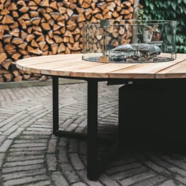 Mesa de fuego Cosiloft 120 redonda negra con cristal en una terraza de adoquines grises y de fondo una leñera llena de troncos
