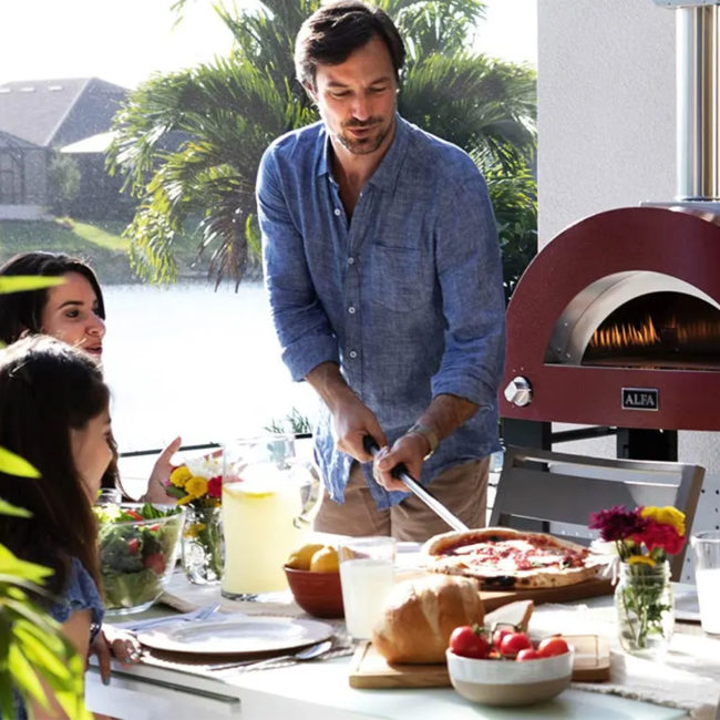 Escena familiar sentadas una mujer y una niña a la mesa mientras el padre sirve una pizza al fondo se ve el Horno MODERNO 3 Pizze Gas encendido de color rojo y sobre su base.