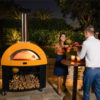 Horno MODERNO 5 Pizze Gas Amarillo encendido y en su base que hace también de leñera. Está en una terraza delante de una jardinera de piedra A la derecha se ve una pareja comiendo pizza en una mesa alta Es una escena nocturna vista lateral