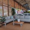 Conjunto jardin Country M de 4 Seasons Outdoor. Se trata de una rinconera de aluminio en gris claro y amplios cojines en el mismo tono con mesitas auxiliares de teca a cada extremo. Además las patas de la mesa y del sofá rinconera son muy originales creando un rectángulo que corta los tablones quedando a la misma altura y a la vista. Al igual pasa con las patas de la mesa que van en sentido inverso a los listones de teca creando un diseño muy original. Está colocado dentro de la exposición de De Tropen.