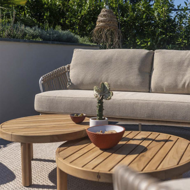 FINN center table set from 4 Seasons Outdoor. Two very attractive tables, completely made of teak. The Ø80cm table has a height of 30cm and the Ø60cm table has a height of 35cm, both with three round legs. Front view, behind it you can see the DALIAS garden sofa and behind it a white planter with many intense green plants.