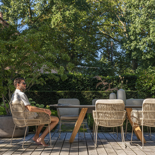 Silla jardín MURCIA de 4 Seasons Outoor® vista oblicua en una terraza de madera se ven 5 sillas al rededor de una mesa y en una hay un hombre sentado con gafas de sol y descalzo