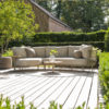 The Sardinia garden sofa by 4SO is a curved sofa with a chaise longue, seating 4. Its curved stainless steel frame is finished in a beautiful tan beige, a rich, toasted shade of café au lait. Rope in the same tones creates a triangular pattern that allows for airflow and adds a touch of sophistication. The generous cushions ensure exceptional comfort. Front view of the sofa with the Volta coffee table in front. Lifestyle photo on a wooden decked terrace surrounded by lush green hedges.