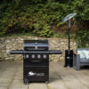 The Fireside Sahara A450 gas barbecue features an enameled cast iron grill that seals in flavours and 4 stainless steel burners plus a side burner. A thermostat in the lid allows you to regulate and monitor the temperature while grilling. This barbecue is a worthwhile investment in quality. It has an integrated and easy-to-use electric ignition system. It features a concealed compartment for the gas cylinder. A wooden cutting board extends from the right side shelf. The knobs and handles are silver, while the rest is black. An elegant combination that never fails. Front view of the barbecue closed on a terrace with grey tiled flooring, with a stone wall and many plants visible behind it in the background.