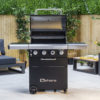 The Fireside Sahara A450 gas barbecue features an enameled cast iron grill that seals in flavours and 4 stainless steel burners plus a side burner. A thermostat in the lid allows you to regulate and monitor the temperature while grilling. This barbecue is a worthwhile investment in quality. It has an integrated and easy-to-use electric ignition system. It features a concealed compartment for the gas cylinder. A wooden cutting board extends from the right side shelf. The knobs and handles are silver, while the rest is black. An elegant combination that never fails. Front view of the Sahara A450 gas barbecue open on a terrace with grey tiled flooring, with a wooden fence and several plant pots visible in the background.
