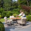 Outdoor dining set consisting of the Saba Low 130cm table and 4 Sardinia Low chairs around it. Lifestyle photo showing a young woman placing two vases in the centre of the table, on a terrace with grey flooring surrounded by gardens, with trees of different shades visible in the background. Find it at De Tropen, official supplier of 4 Seasons Outdoor in Spain.