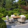 Outdoor dining set consisting of the Saba Low 130cm table and 4 Sardinia Low chairs around it. Lifestyle photo showing the table surrounded by 4 chairs on a circular rug in the same latte tone, on a terrace with grey flooring surrounded by gardens, with trees of different shades visible in the background. Find it at De Tropen, official supplier of 4 Seasons Outdoor in Spain.