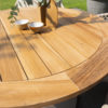 Mesa Saba Baja de 4SO Foto ambiente desde arriba, detalle del tablero de teca de un marrón anaranjado intenso y profundo característico de esta madera tropical natural y certificada FSC. Hecho de listones y terminado con un borde de listones perpendiculares redondeados para darle forma circular al ancho borde. Se ve un poco de la base de la pata hecha de aluminio y terminada en un color latte (café con leche o beige tostado) Color tendencia del 2025. Cómprala en De Tropen, tu experto en muebles de jardín y terraza. Foto frontal un poco desde arriba.