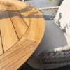 Saba Low Table by 4SO. Photo taken from above, showing a detail of the teak tabletop in a deep, intense orange-brown characteristic of this natural, FSC-certified tropical wood. Made of slats and finished with a rounded, perpendicular slat edge to create a circular shape. The Sardinia low chair, made of stainless steel and finished in a latte colour (coffee with milk or toasted beige), a trend colour for 2025, is visible. Buy it at De Tropen, your expert in garden and patio furniture. Front view taken slightly from above.