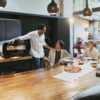 URBE electric oven by Alfa Forni. Three friends gathered around a table with drinks and a pizza, the oven is on the left, closed and lit, lifestyle photo. Find it at De Tropen, official Alfa Forni supplier in Spain and Portugal.