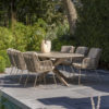 Comedor Prado barrel Fabrice de 4 Seasons Outdoor Foto ambiente del comedor en oblicuo se ve la mesa Prado barrel de tablero cerámico con forma de barril, además las patas cruzadas de acero inoxidable y terminadas en latte. Rodeada de 6 sillas Fabrice hechas de acero inox y terminadas en un color latte (café con leche o beige tostado, color tendencia del 2025). Se embellecen con la cuerda en diferentes tonos de beige anudada a la estructura desde los reposabrazos hasta la base del asiento rodeando toda la silla y creado un patrón característico de la colección Fabrice. Colocado sobre una terraza de losetas grises delante se aprecia el filo de una piscina y detrás está rodeado de vegetación. Cómprala en De Tropen, tu experto en muebles de jardín y terraza.