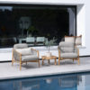 Nora teak armchair set by 4SO. This set of teak armchairs features an ergonomic, rounded shape, embellished with taupe rope knotted to the frame from the armrests to the seat base, encircling the entire chair and creating a pattern characteristic of the Nora collection. Both the legs and the frame are cylindrical and smooth. The set includes both seat and back cushions in different shades of toasted beige. Buy it at De Tropen, your expert in garden and patio furniture. The photo shows the Nora armchair set (2 armchairs) with the Finn table in the middle, placed on a concrete terrace in front of a house where the interior living room is visible through a large glass window. In front of the armchairs, there is a swimming pool reflecting the set.