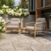 Nora teak armchair set by 4SO. This set of teak armchairs features an ergonomic, rounded shape, embellished with taupe rope knotted to the frame from the armrests to the seat base, encircling the entire chair and creating a pattern characteristic of the Nora collection. Both the legs and the frame are cylindrical and smooth. The set includes both seat and back cushions in different shades of toasted beige. Buy it at De Tropen, your expert in garden and patio furniture. The photo shows the Nora armchair set (2 armchairs) with the Finn table in the middle, placed on a tiled terrace in front of a modern wooden house with a metal railing. In the foreground to the left, you can see the grass, and behind, beautiful white hydrangeas cascade over the side wall.