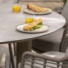 Donato garden table by 4SO. Made of aluminium finished with latte-coloured powder coating, a trendy café-au-lait or toasted beige tone, and tempered glass printed with ceramic motifs in grey tones. Its 3 cylindrical legs characterise this outdoor table, with the Lazy Susan rotating tray in the centre, finished in the same printed tempered glass. Find it at De Tropen, official supplier of 4 Seasons Outdoor in Spain. Side view detail of the table with Fabrice chairs in latte around it, with two plates and glasses on the table, each with a sandwich and orange juice.