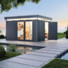 Foto ambiente Caseta CasaNova en gris oscuro de Biohort, se ve la caseta con doble puerta abierta y a través de los elementos de cristal instalados en el lateral, se ve a una pareja en toalla con la sauna detrás. Instalada sobre una terraza de piedra junto a una piscina natural y un camino del mismo tipo de suelo y rodeado de césped muy verde. Amplía tus horizontes con De Tropen®, ven a descubrir las posibilidades de personalización de la CasaNova. En la mayor tienda de muebles de jardín de la Costa del Sol.