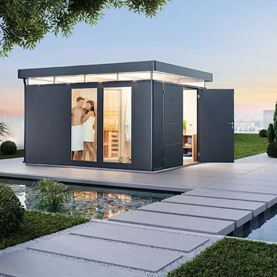 Foto ambiente Caseta CasaNova en gris oscuro de Biohort, se ve la caseta con doble puerta abierta y a través de los elementos de cristal instalados en el lateral, se ve a una pareja en toalla con la sauna detrás. Instalada sobre una terraza de piedra junto a una piscina natural y un camino del mismo tipo de suelo y rodeado de césped muy verde. Amplía tus horizontes con De Tropen®, ven a descubrir las posibilidades de personalización de la CasaNova. En la mayor tienda de muebles de jardín de la Costa del Sol.