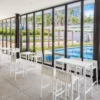 Photo of the Spritz garden stool by Vondom. Featuring the characteristic slat pattern of the Spritz series. Several combinations are shown: two high tables with four stools each and one table with two stools, all in white, in a glass-enclosed room overlooking a large pool and garden. Buy it online at De Tropen, the largest garden furniture store in Malaga.